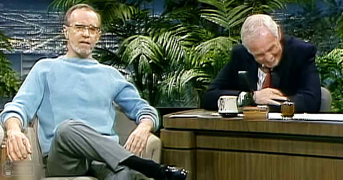 An older adult male wearing a light blue sweater and gray trousers is seated at a round wooden desk in what appears to be an indoor studio set for a talk show or interview, with artificial green foliage in the background. The setting includes bright, even lighting suggesting daytime.
