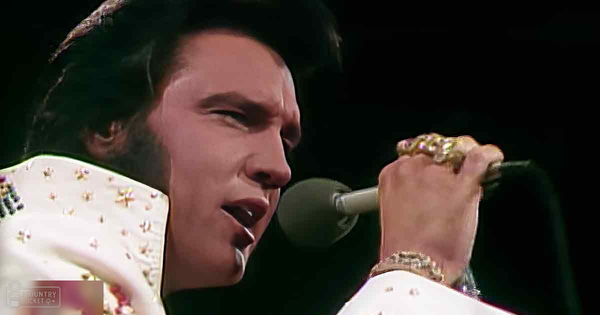 Adult male performer in a white suit jacket adorned with intricate beadwork and sequins, holding a microphone on stage under focused lighting.