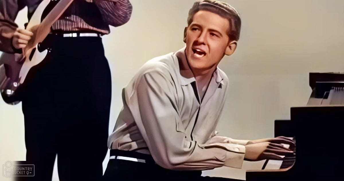 A young adult male sings and plays the piano in a plain studio setting with even lighting.
