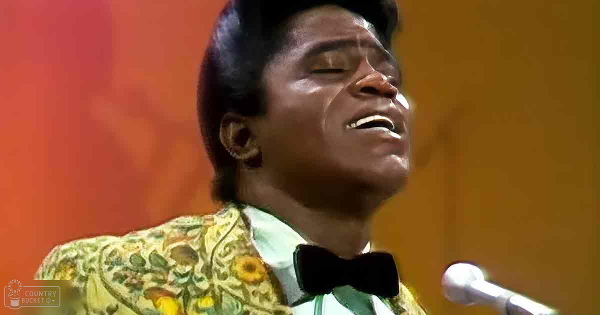 An adult male singer wearing a patterned green, yellow, and brown floral jacket with a high collar and white shirt with a bow tie sings into a microphone in what appears to be a recording studio setting.