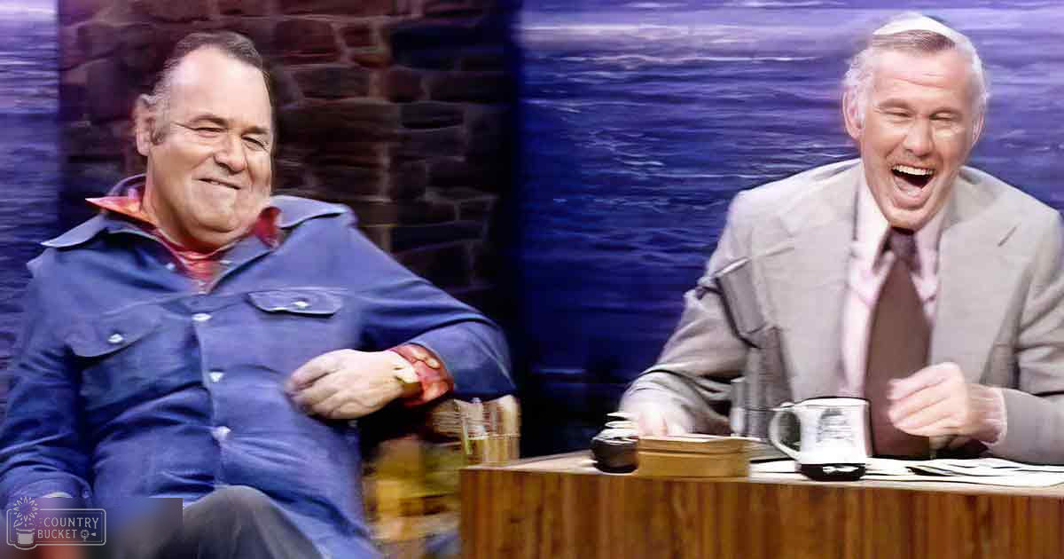 Talk show setting with two middle-aged men, one wearing a blue denim jacket and red shirt, another in a light gray suit with pink checkered shirt, both indoors on what looks like a studio set.