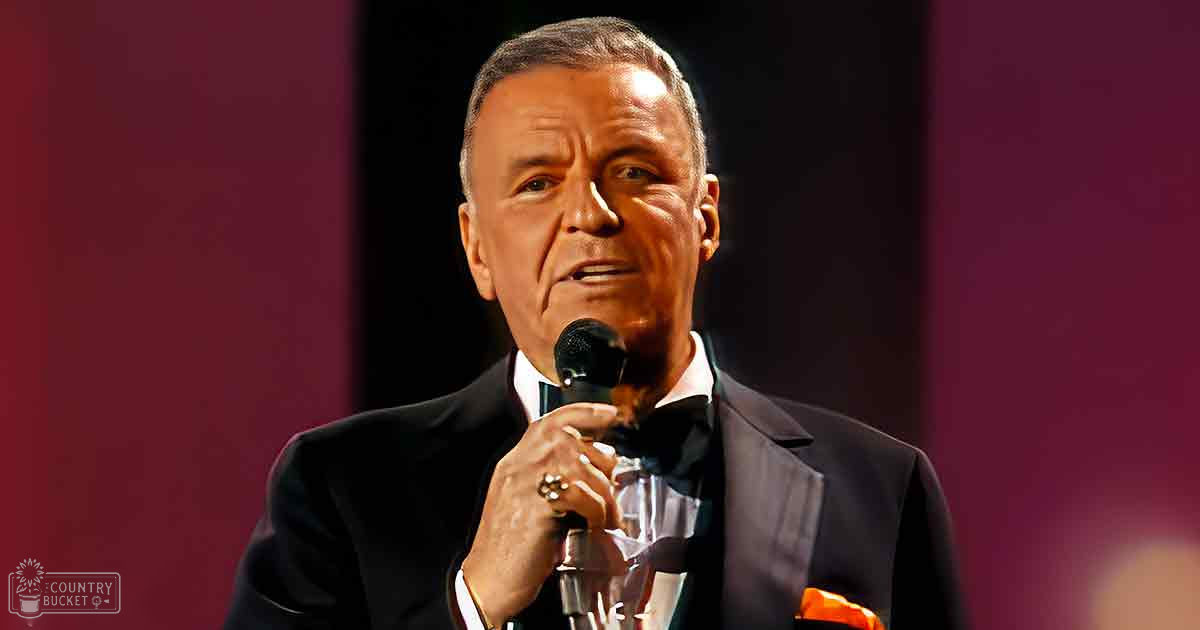 A middle-aged adult male in a tailored tuxedo with a bow tie, holding a microphone against an indoor backdrop with stage lighting.