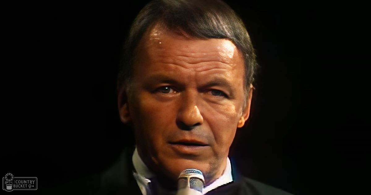 A middle-aged adult male in a formal suit jacket and collared shirt is speaking indoors under focused stage lighting.