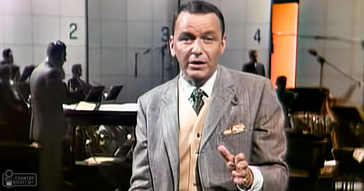 An adult male wearing a gray pinstripe suit jacket, light-colored vest, white shirt, patterned tie, pocket square, sitting at an office desk with numbered cubicles in the background during daytime.