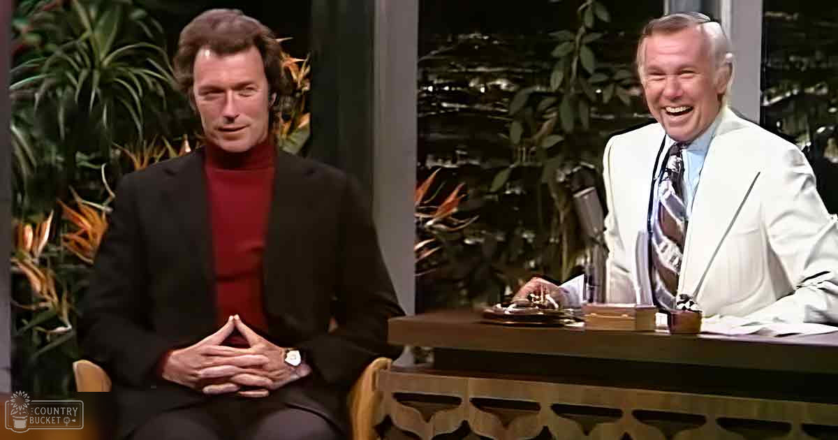 A talk show host wearing a dark suit jacket over a red turtleneck sweater seated at a desk with another older adult male in a white blazer, surrounded by studio furniture and greenery.