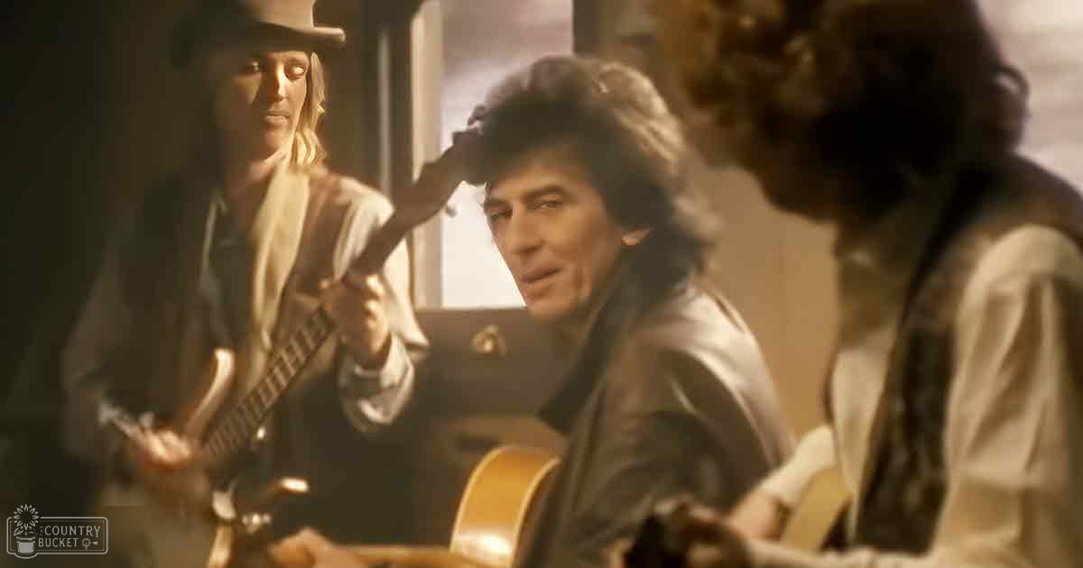 Middle-aged man with dark hair wearing a leather jacket seated inside what appears to be an interior setting resembling a train car, surrounded by musicians playing guitars.