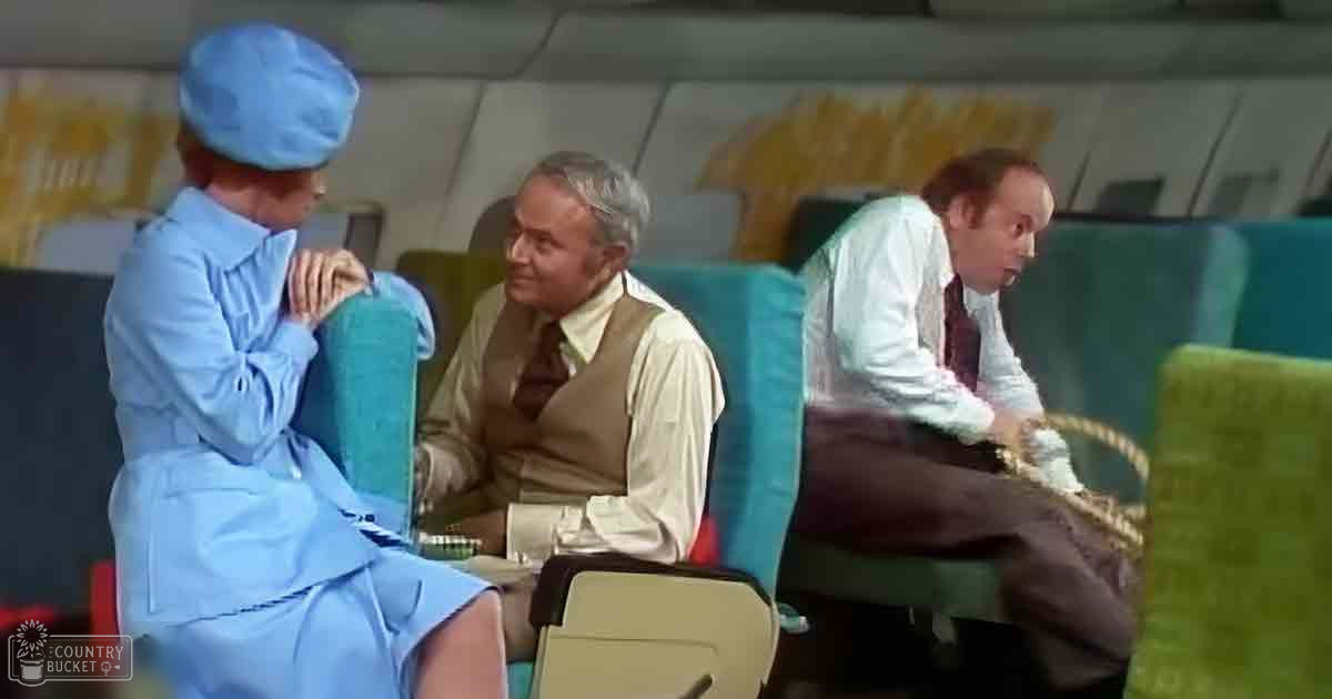 Flight attendant in a light blue uniform with hat, seated among passengers inside an airplane cabin, wearing various clothing items and holding a woven basket.