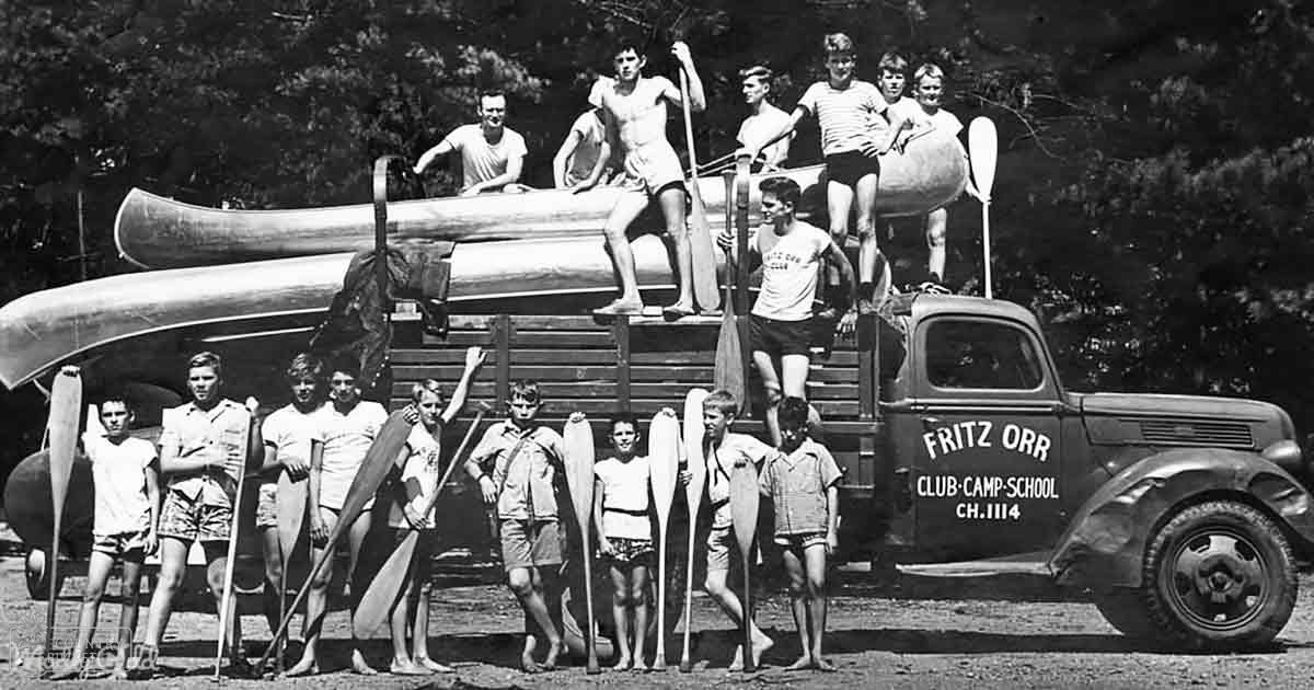 Children in shorts and T-shirts at Fritz Orr Club Camp School with vintage pickup truck labeled CH.1114, canoes on top, outdoors during summer day.