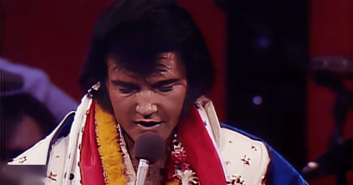An adult male performer wearing a white and gold costume with intricate designs, including a lei around his neck, holds a microphone on stage under spotlight effects.