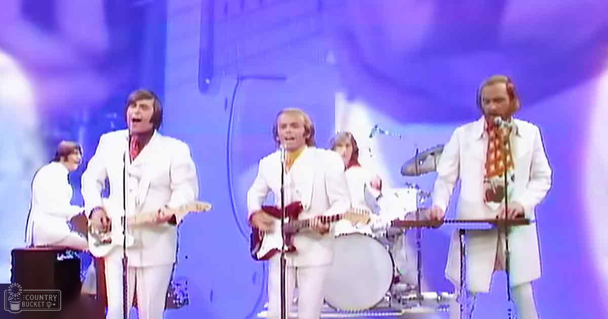 Adult male musicians in white suits performing on a stage with electric guitars, drums, and keyboard under bright stage lights.
