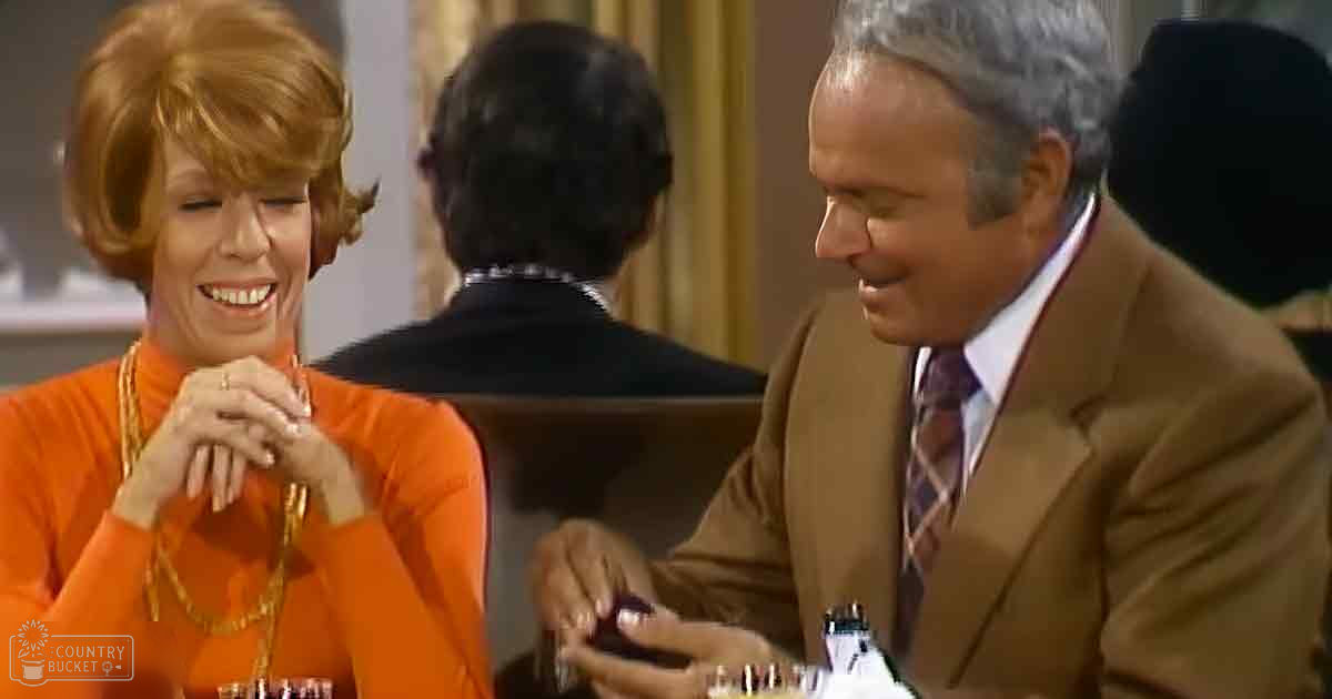 A couple seated at a table indoors, possibly in a restaurant, with warm ambient lighting; woman wears an orange long-sleeved top with gold necklace, man in brown suit jacket and patterned tie.