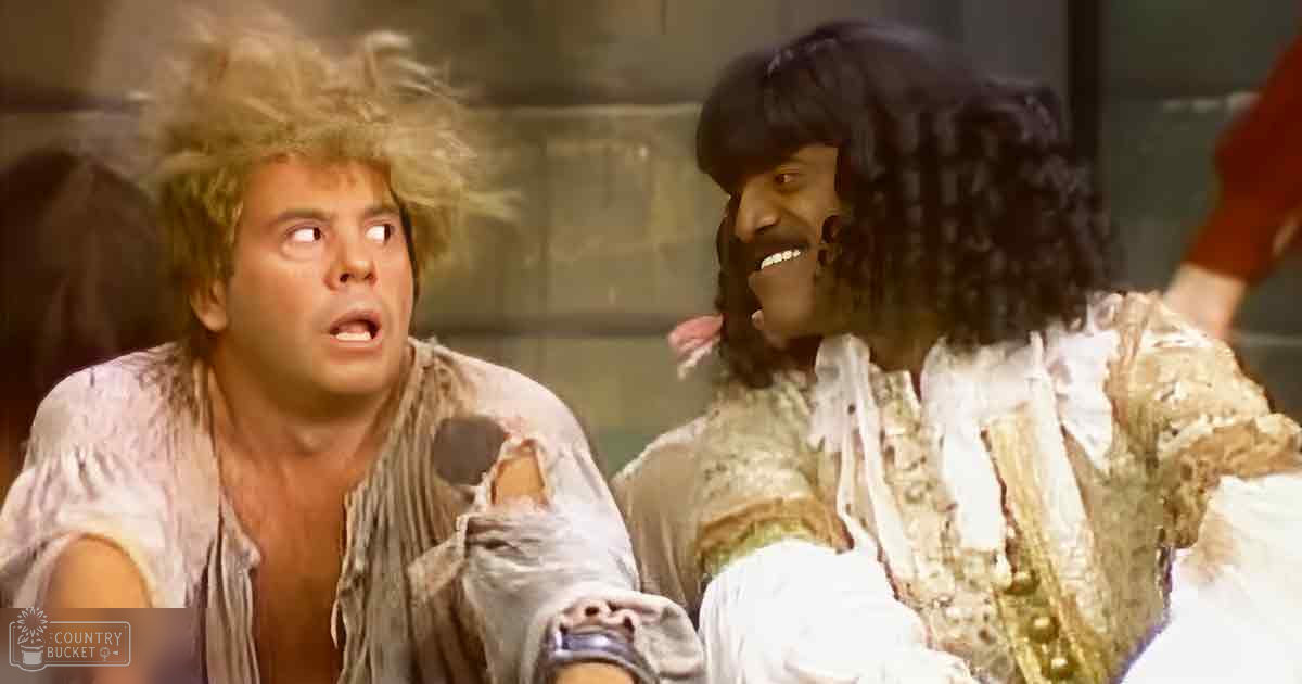 Two adults dressed in tattered white and gold costumes stand against a dark stage backdrop, suggesting a theatrical production.