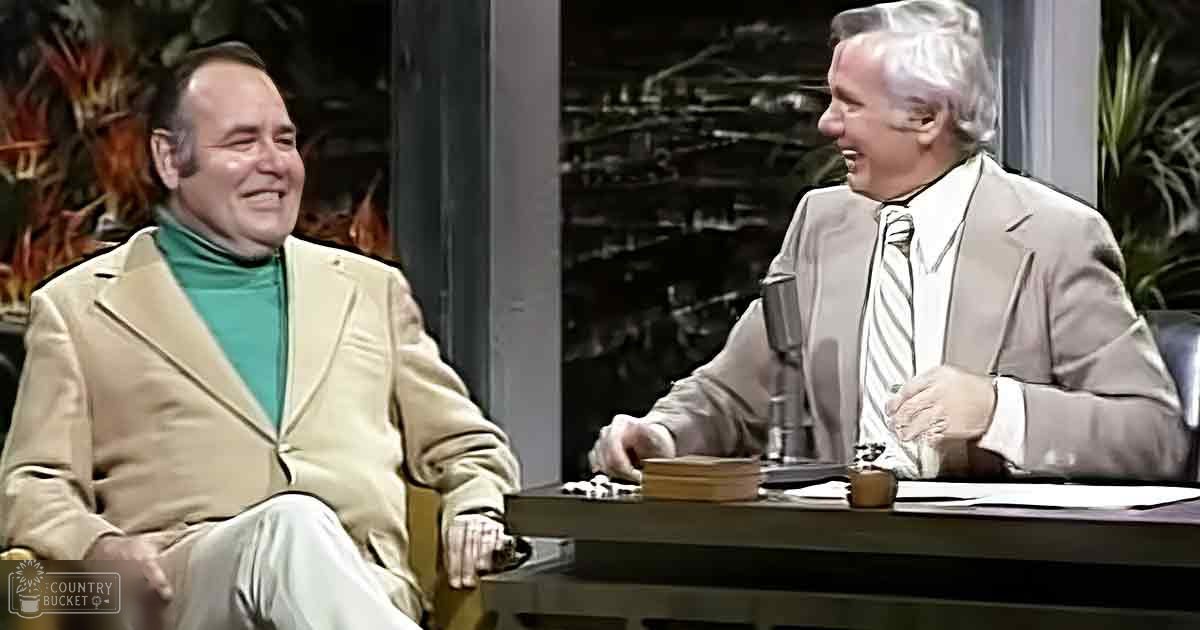 Talk show setting with two middle-aged men in an indoor studio; beige blazer over a green turtleneck sweater, light brown suit jacket and white shirt with vertical stripes. Plants and reflections suggest daytime but not sunny.
