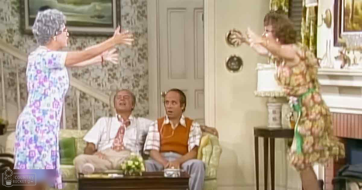A sitcom scene set indoors in a living room with green couches, patterned cushions, wooden furniture, and various home decor items.