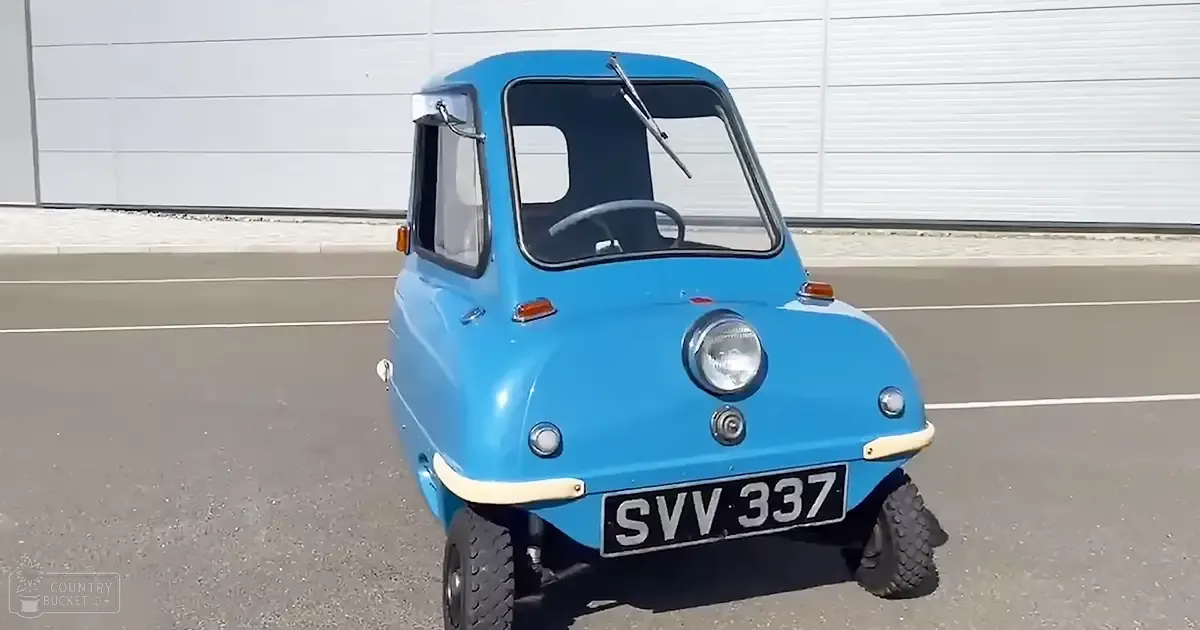 A small, compact blue car with rounded edges and a vintage design sits outdoors on a paved road against a white wall under clear daylight.