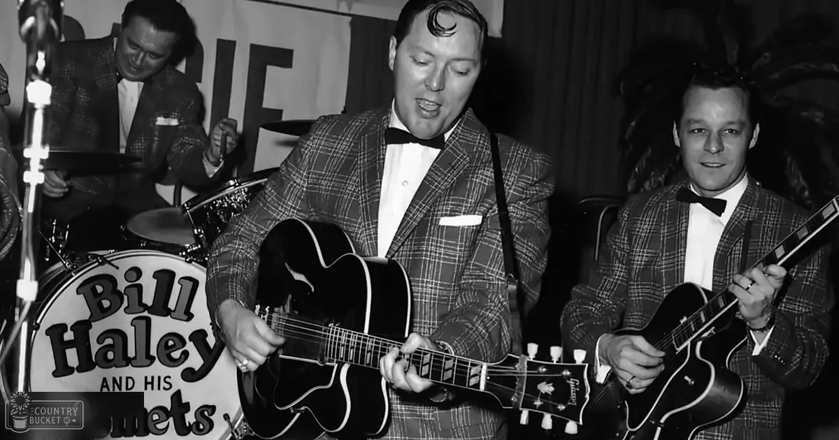 A performer wearing a plaid suit jacket, white shirt, and bow tie at what appears to be a concert or performance setting with an acoustic guitar and drum set in the background.