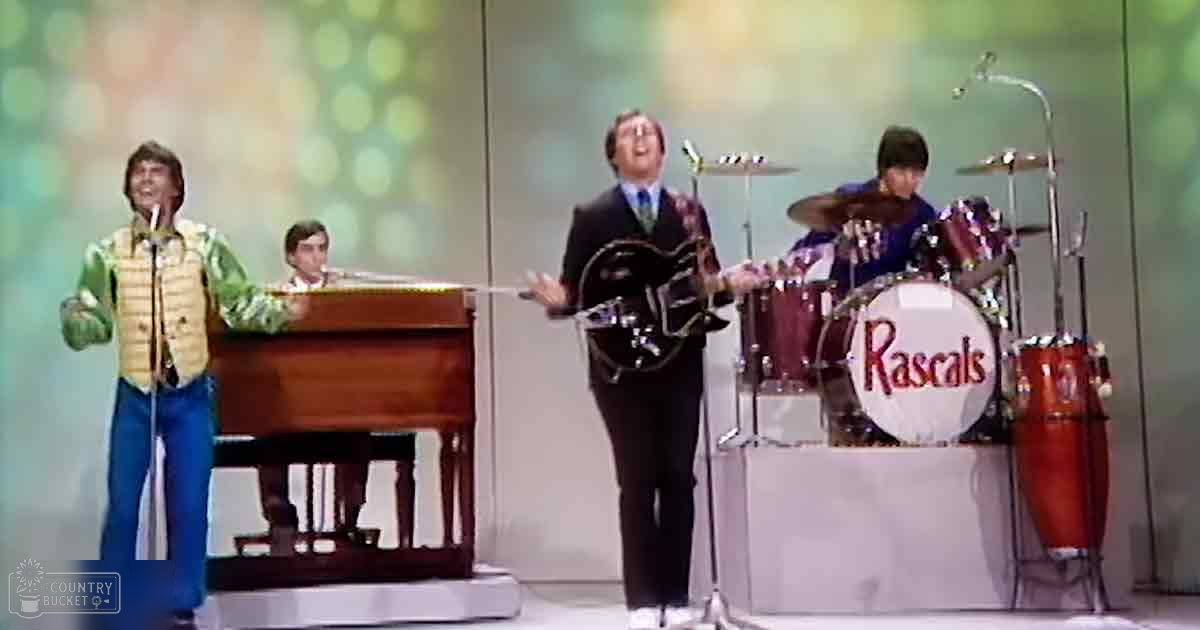 A lively musical performance with 'The Rascals' in an indoor studio setting, with performers dressed in mid-20th-century attire and a grand piano as the centerpiece.