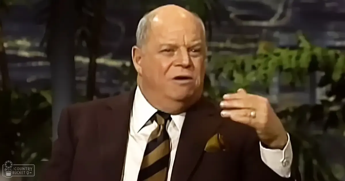 An older adult male seated indoors against a backdrop of blurred greenery, wearing a dark suit jacket with a white shirt and striped tie, in a studio setting from the mid-20th century.
