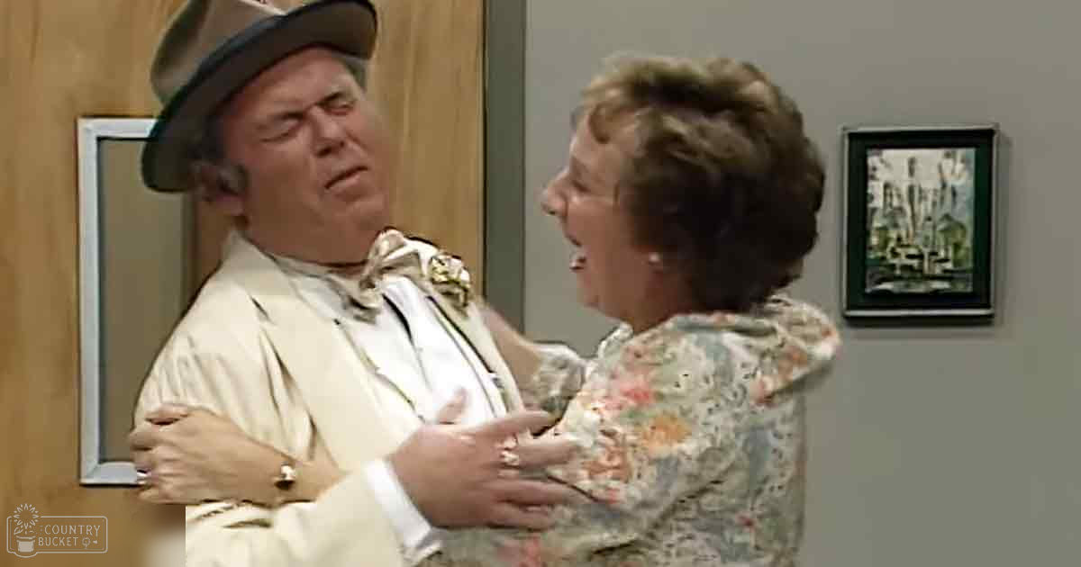 An adult male wearing a light-colored suit, white shirt, possibly with a bow tie, dark hat, and ring on his left hand stands indoors alongside an adult female in a floral-patterned blouse. Framed artwork and a wooden door are visible in the background.