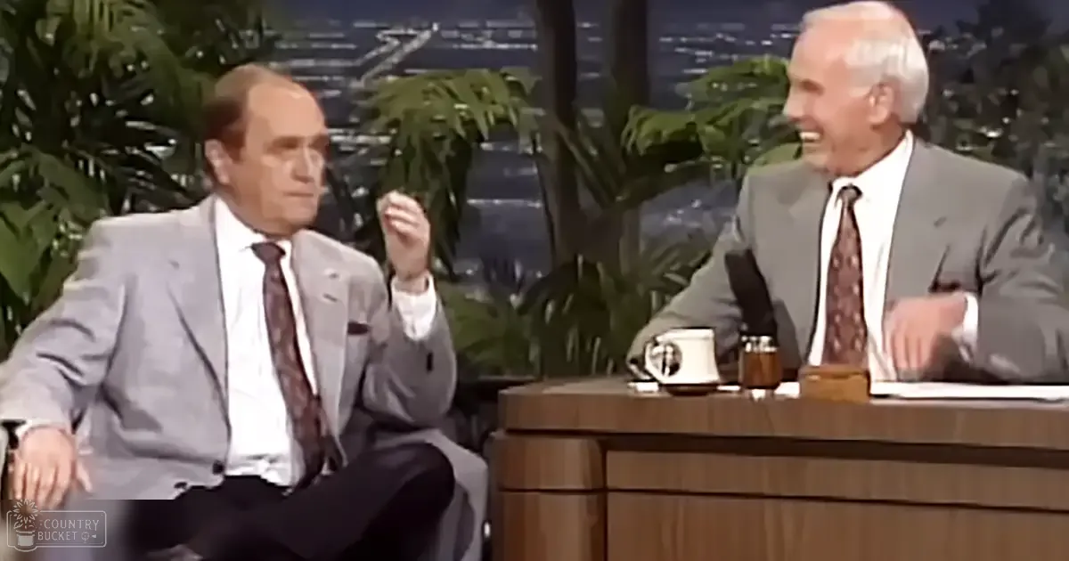 Two older adult males dressed in formal gray suit jackets with white shirts and dark patterned ties, seated at a wooden desk with a mug, container, and notebook in an indoor studio setting with large green plants and a cityscape or water view.