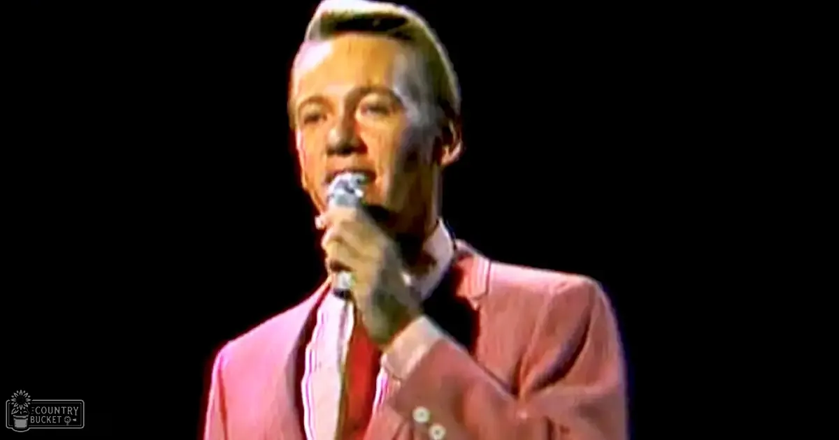 A middle-aged male performer in a vintage pink suit with white stripes, holding a microphone against a dark background during an indoor performance.