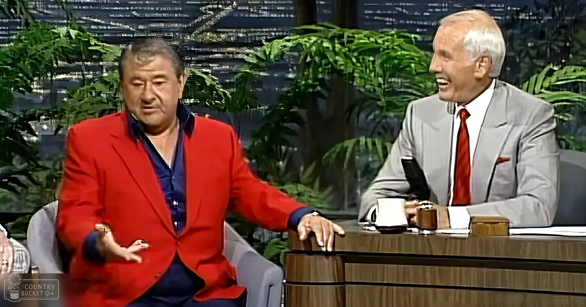 Adult male and older adult male seated at a television talk show set with cityscape and greenery background, daytime lighting.