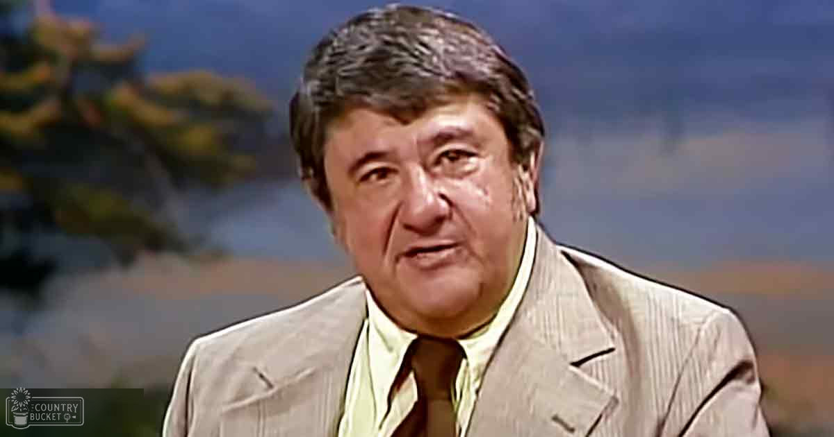 An older adult male wearing a light-colored suit jacket with subtle pinstripes, a light green shirt, and a dark tie, set against a blurred backdrop suggesting an Asian-themed studio environment.