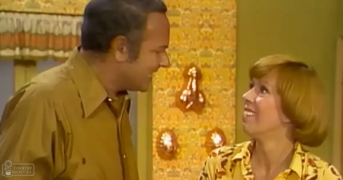A man wearing a mustard-yellow button-up shirt, an adult male singer; a woman in a yellow patterned blouse with ruffled collar, an adult female singer, indoors with mid-20th century decor and stylized wallpaper.