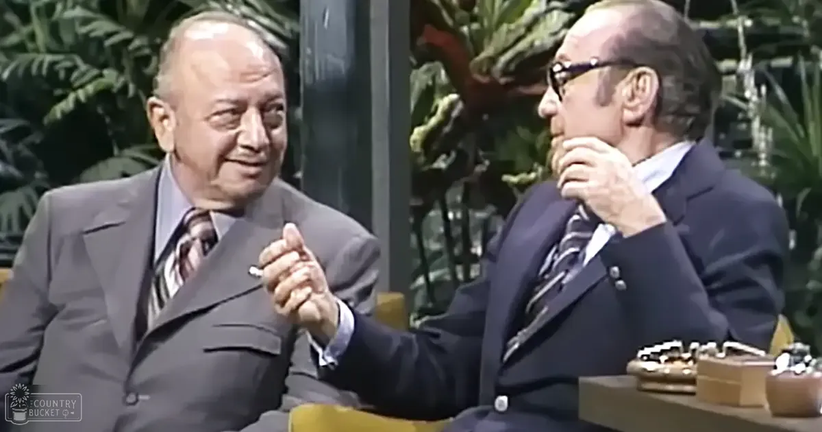 Two older adult males in formal attire seated against a backdrop with greenery, suggesting an indoor studio environment for a talk show or interview.