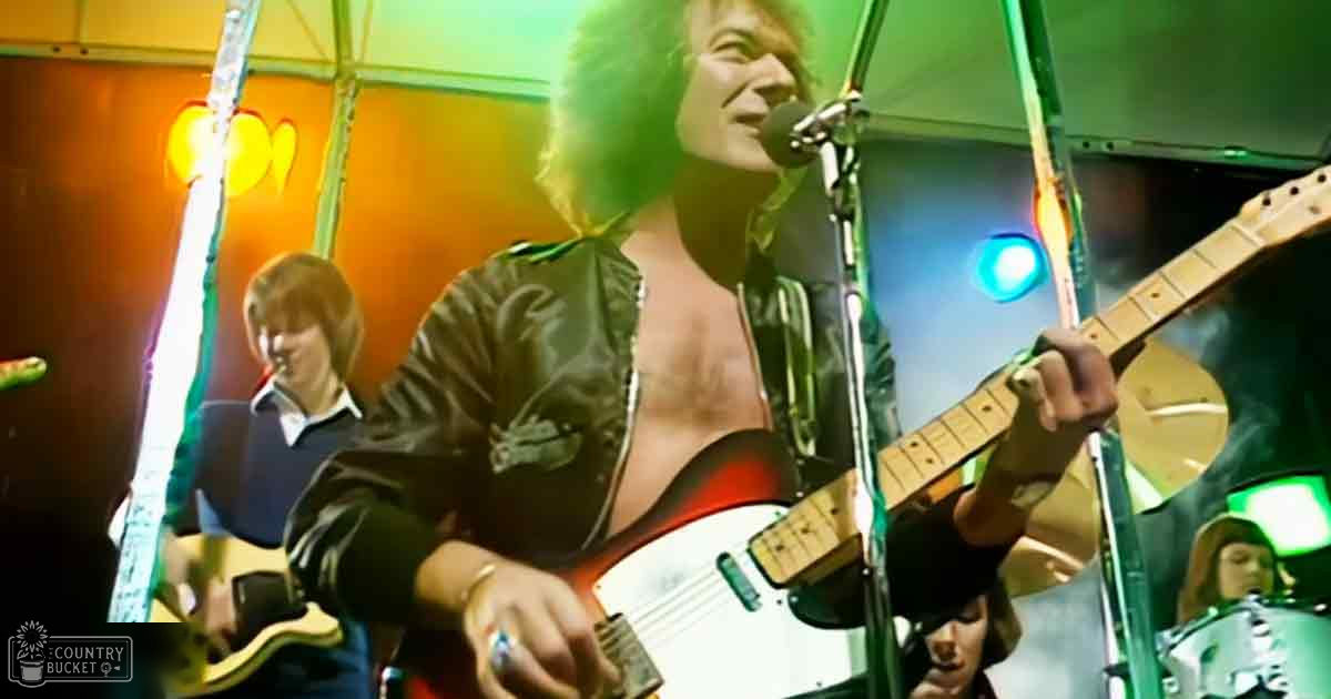 A young adult male performing live music, wearing a dark jacket with shiny material, surrounded by stage lights in an indoor venue.