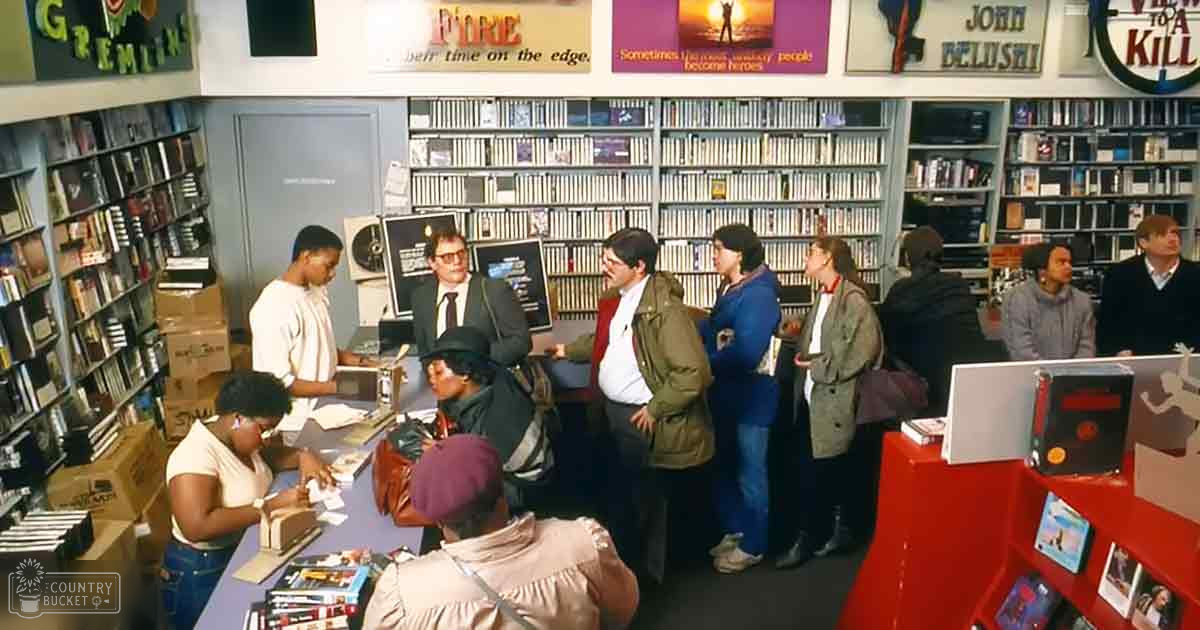 A group of people in a video rental store, including an adult male wearing glasses and a suit jacket behind the counter.