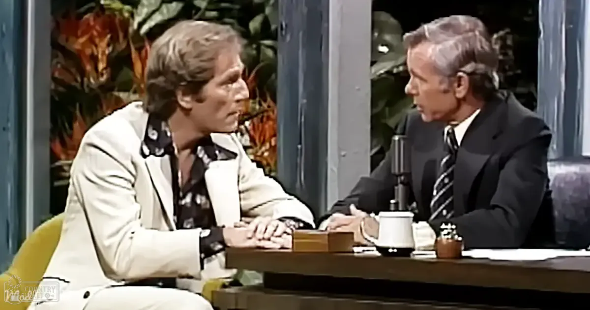 Two individuals seated at a talk show table with wooden furniture and indoor plants, suggesting an indoor setting.