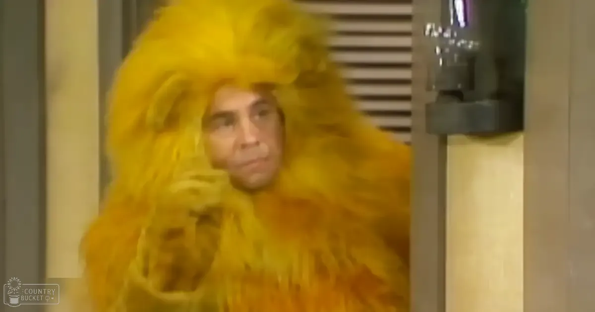 A middle-aged male in a large, fluffy yellow wig and feather-like costume at an indoor event or performance setting.