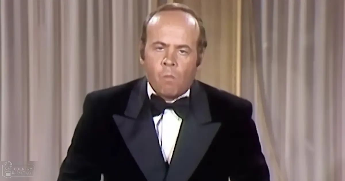 An older adult male in a black tuxedo with a white dress shirt and a black bow tie, standing against light-colored curtains indoors.