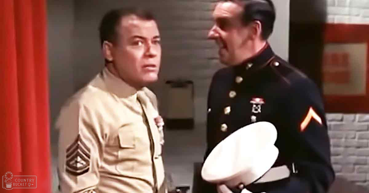 Two adult males in military uniforms, one beige and mid-20th century style, the other dark with insignia, indoors against a red curtain and brick wall. Daytime natural light.