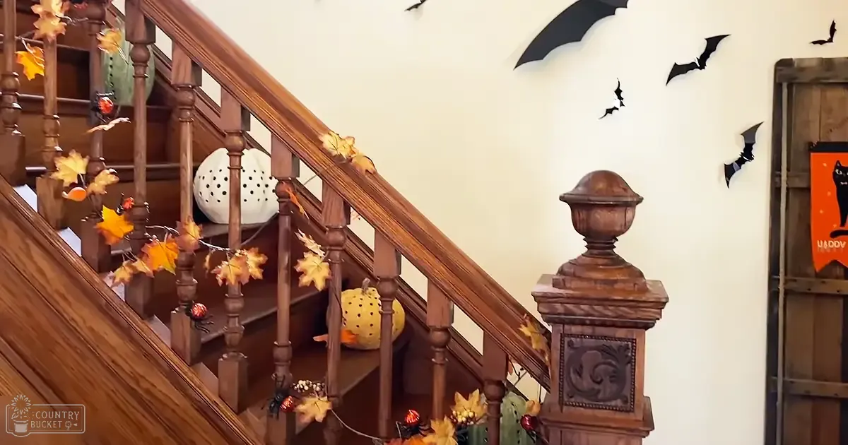 Halloween-themed indoor scene with autumn leaves, pumpkins, and spiders adorning a wooden staircase; bats cutouts on the wall above the stairs.