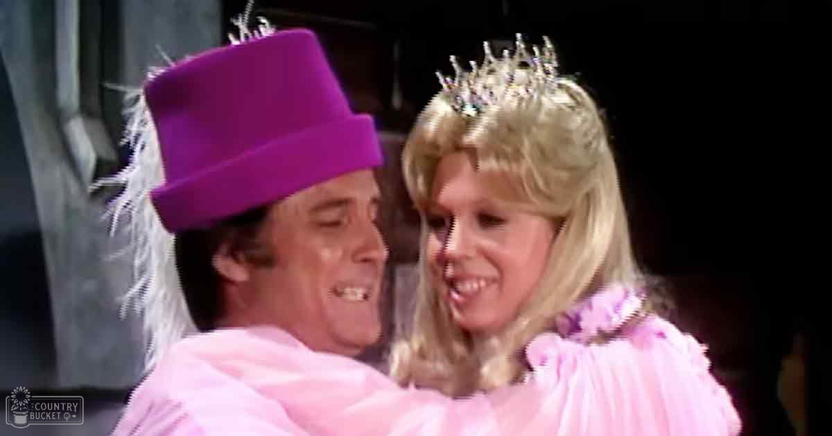 An adult male singer wearing a bright purple top hat with feathers and a pink coat with ruffles, alongside an adult female in a fluffy pink garment adorned with jewels or rhinestones, set against the backdrop of what appears to be a stage or performance room.