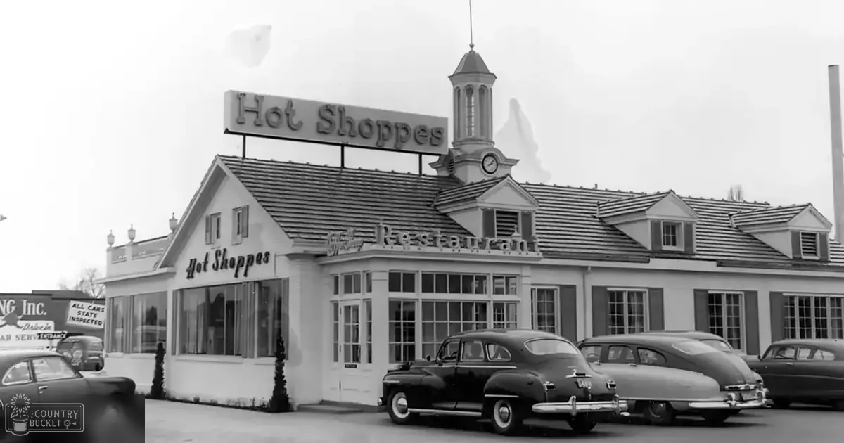 Vintage cars parked in front of a 'Hot Shoppes' restaurant on a commercial street with bare trees suggesting spring or fall, soft daylight casting shadows.
