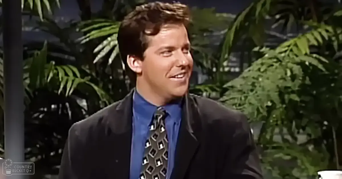 Young adult male in a formal suit jacket and collared shirt with geometric tie, standing indoors with tropical foliage visible through large windows.