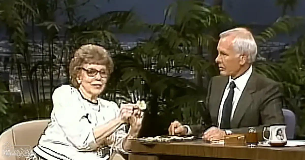 Talk show set with two adults seated at a round wooden table; palm trees in the background suggest an outdoor setting likely near a beach or tropical location, under bright daytime light.