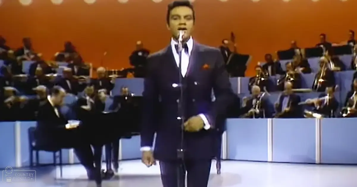 An adult male singer performing on stage with an orchestra in the background, likely at a concert hall under indoor lighting.