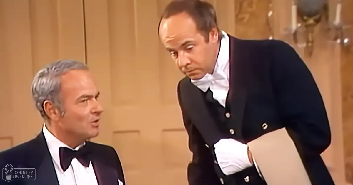 Elegant indoor scene from a theatrical performance or film set with two middle-aged to older adult men in formal attire; ornate door and candlesticks add sophistication.