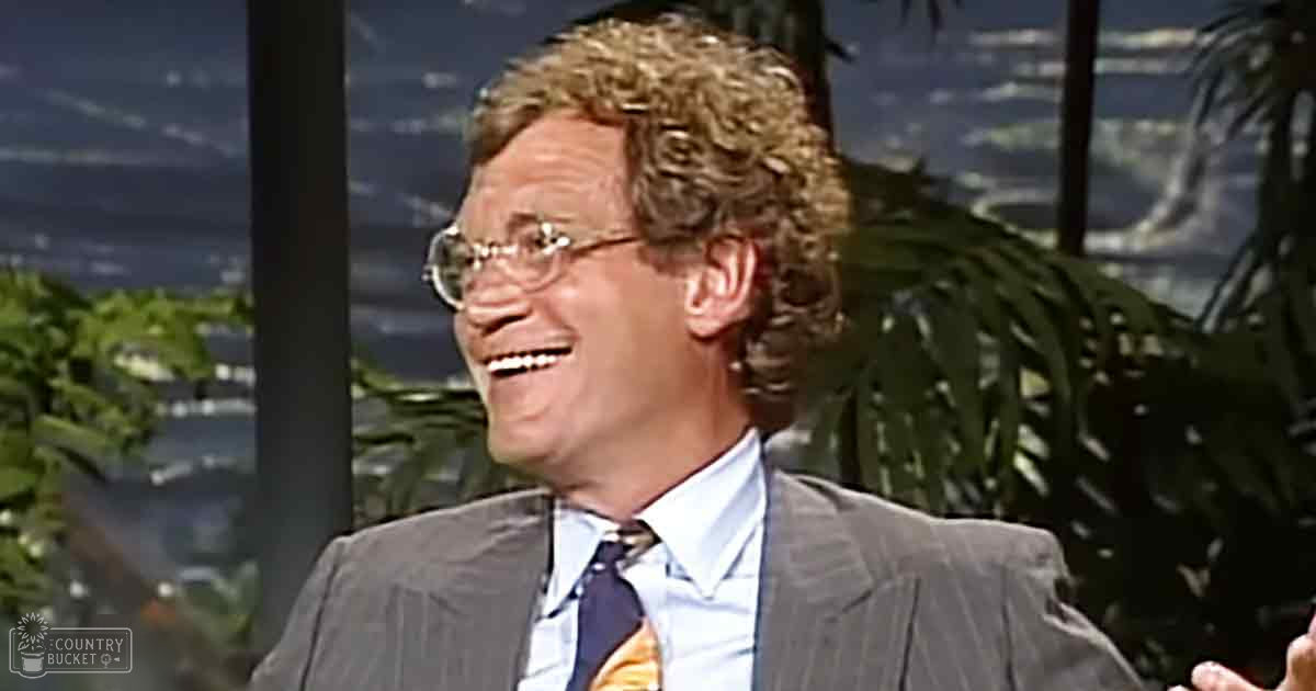 A middle-aged man wearing a gray suit jacket, white dress shirt, and patterned tie sits indoors against a scenic backdrop with large windows showing natural scenery. The lighting is bright and even, typical of television studio settings.