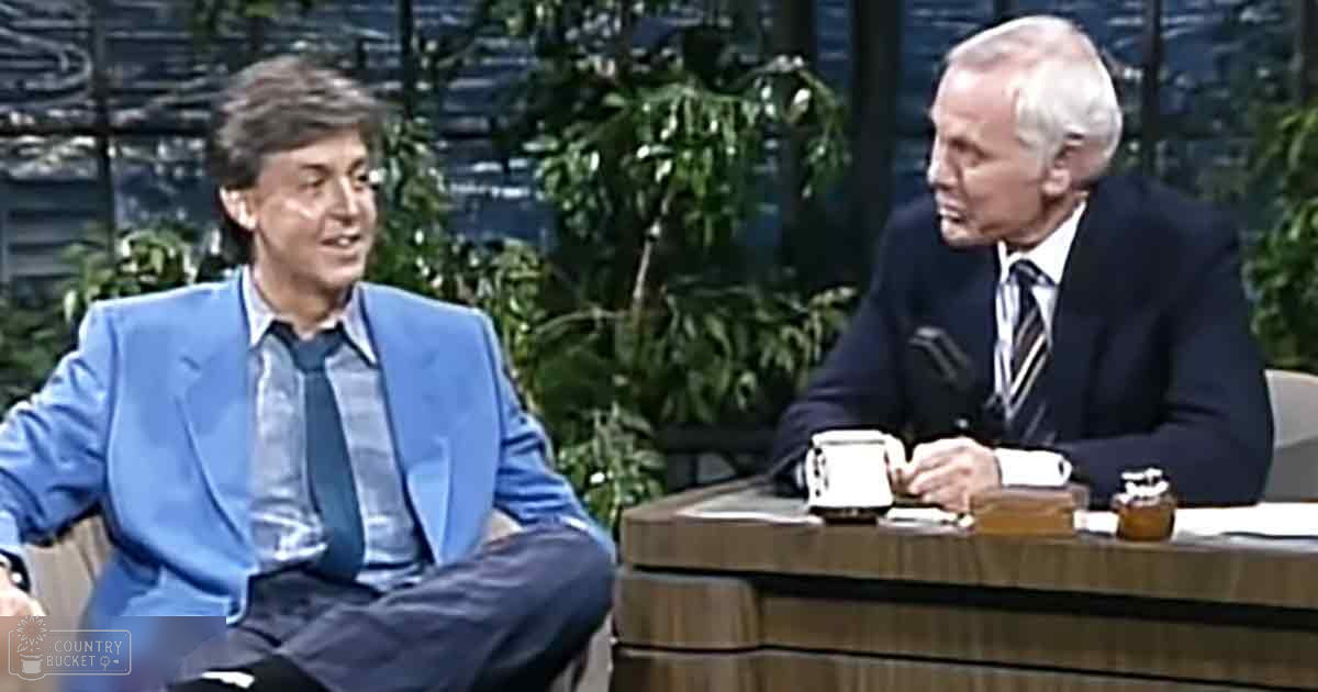 A middle-aged male talk show host in a bright blue suit jacket interviews an older adult male guest wearing a dark suit, set against a backdrop of greenery and indoor studio furniture.