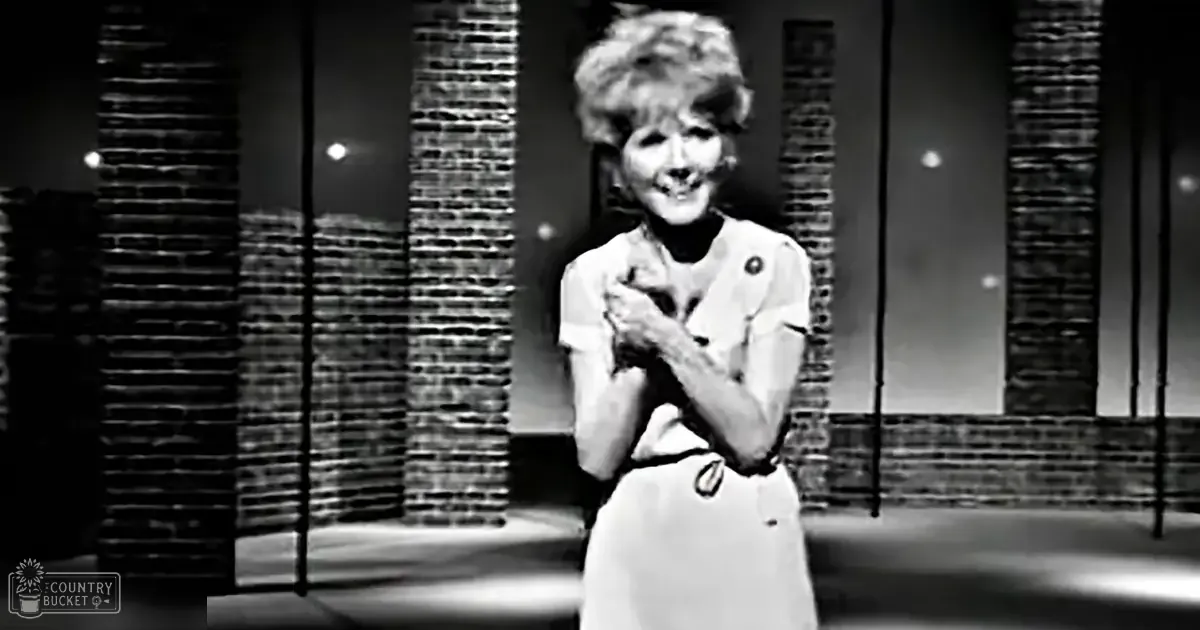 An older adult woman in a performance setting, wearing a vintage-style short-sleeved dress with brick columns and lights suggesting an urban or stage environment.