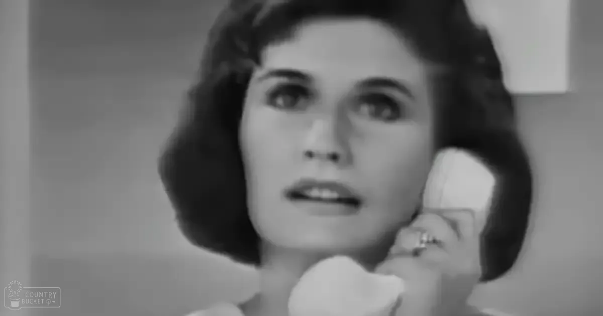 A young adult female wearing a simple, light-colored blouse or dress with short sleeves, holding a telephone near her ear in an indoor setting with minimalistic neutral-toned walls and even lighting suggesting daytime.