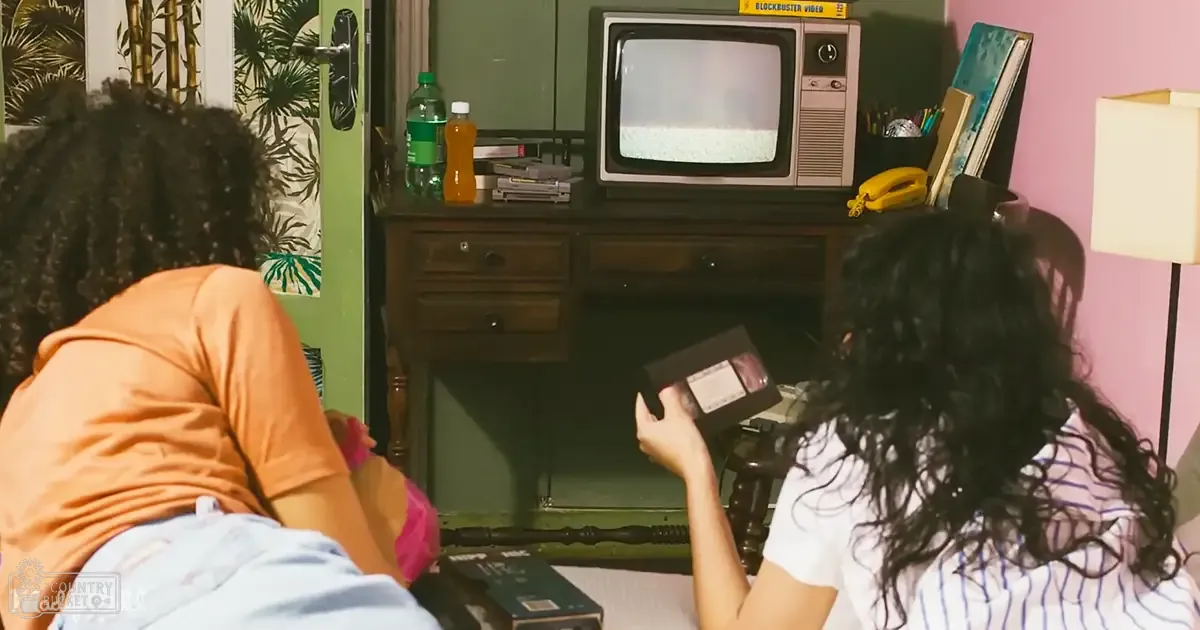 Two individuals in an indoor setting with furniture and home decor; one wearing a peach t-shirt and jeans, another in a striped white shirt.