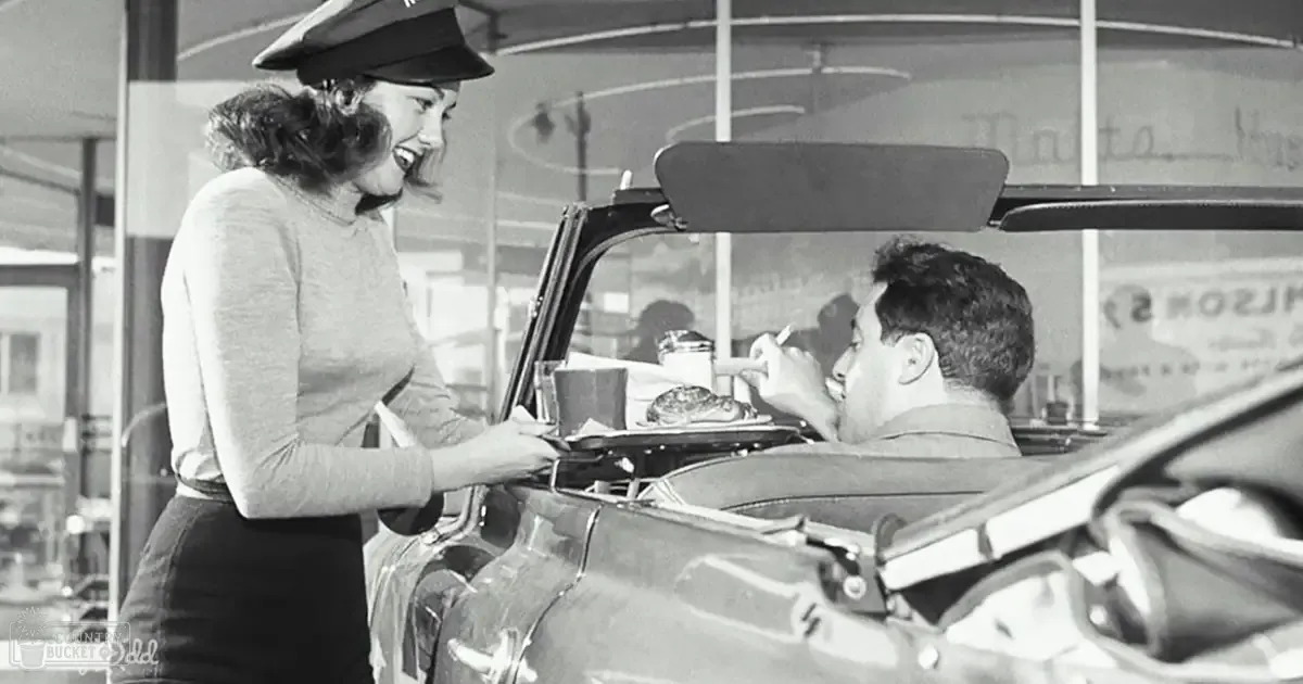 A woman serving food from a tray to a man in his car at a vintage drive-in restaurant, wearing a dark skirt, long-sleeved sweater with ruffled cuffs, and a wide-brimmed hat; the man is eating from a tray.