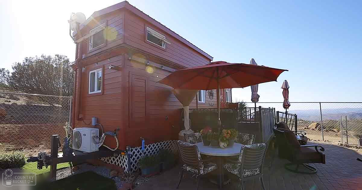 A small wooden house with a patio area, surrounded by potted plants under a clear blue sky during late afternoon or early evening.