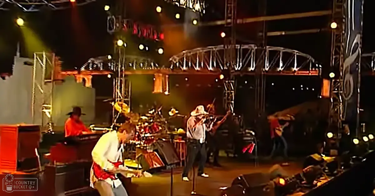 Musical performance at an outdoor concert venue with performers dressed in red and white shirts playing guitars and a violin under artificial stage lighting.
